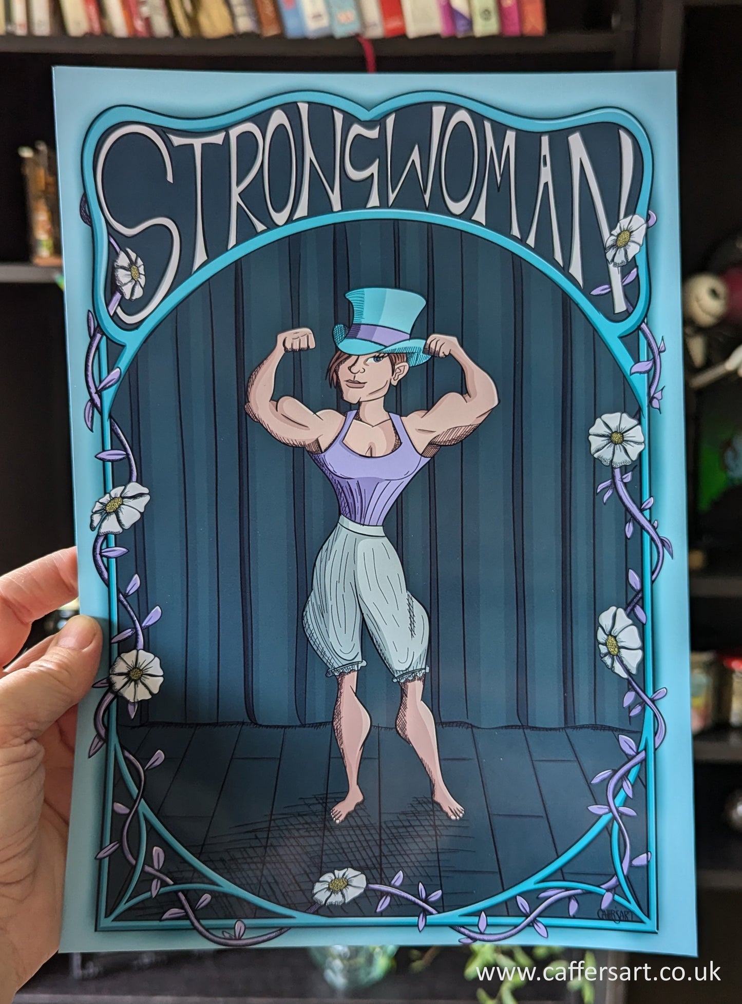 A muscly Victorian strongman doing a double bicep pose. She's wearing pantaloons a lilac corset and a turquoise top hat. There is Strongwoman test along the top and vines and flowers interweaved into the illustrated frame surrounding the print.